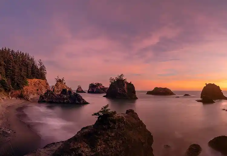 Sea stacks rising from calm water beneath a colorful sunset at Secret Beach on the Oregon coast.