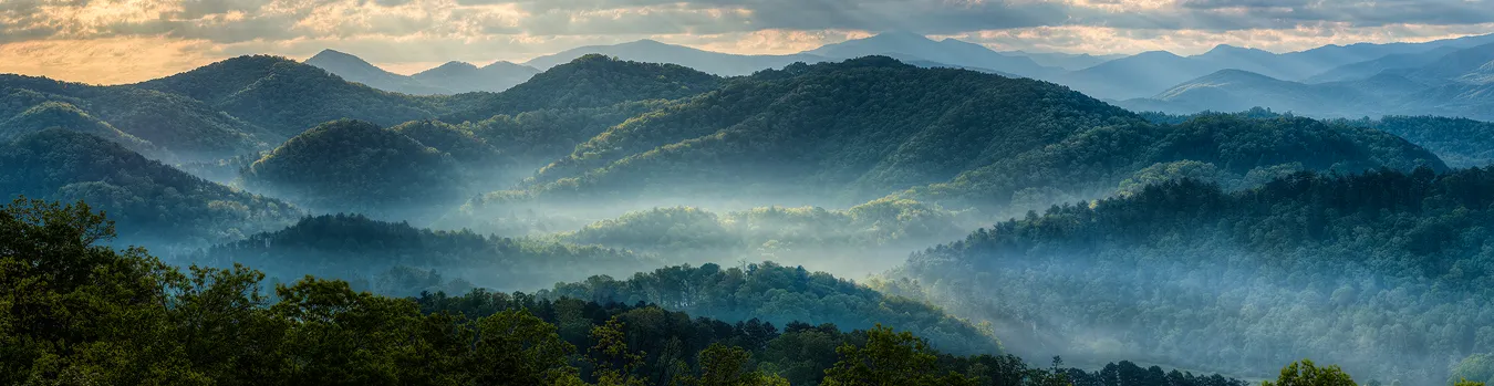 Workshop location; Morning, panorama of the eastern view from an overlook on Foothills Parkway near Great Smoky Mountains National Park. The rolling, forested hills and valleys seen here are a favorite with photographers. In this Spring image the mountains are back lit and a wisp of fog meanders through the valleys. Where the sun breaks through the partly cloudy sky and hits the thin valley fog, it glows and powerful attracts the eye.